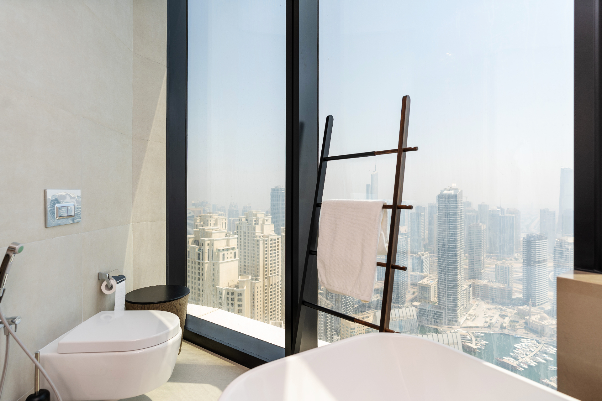 Primary bathroom fragment — wall-hung WC and leaning wooden towel ladder framed by full-height windows onto Dubai Marina