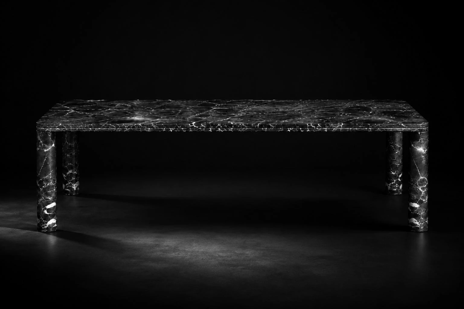 Black-and-white photograph of the Luna Dining Table, a veined stone dining table with cylindrical column legs, set against a black background.