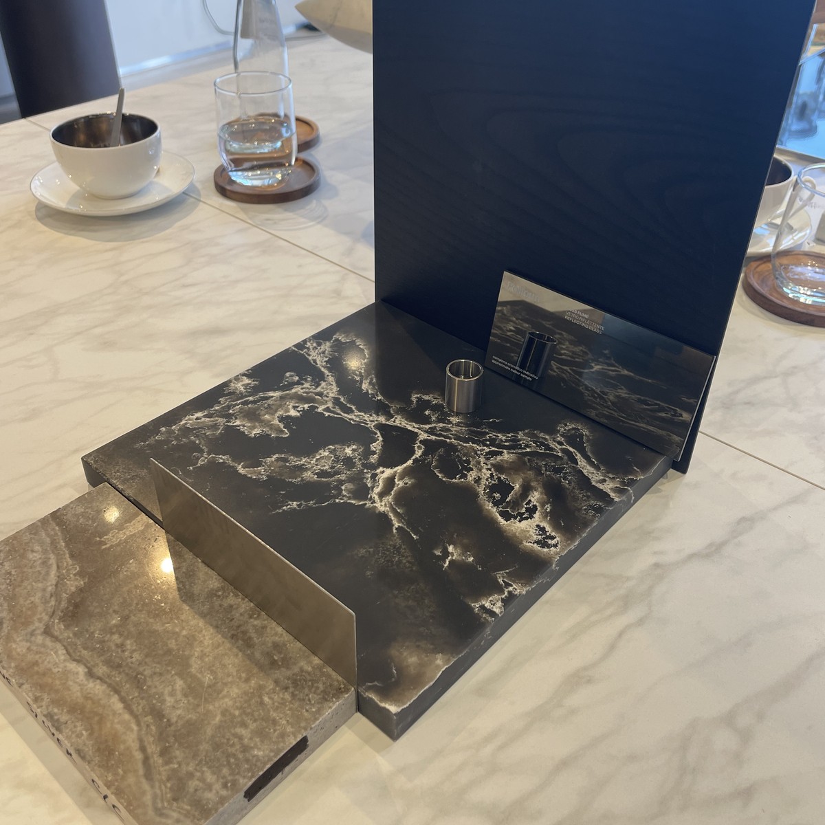 Dark veined marble, travertine and brushed metal samples presented on a pale marble table beside coffee and water.
