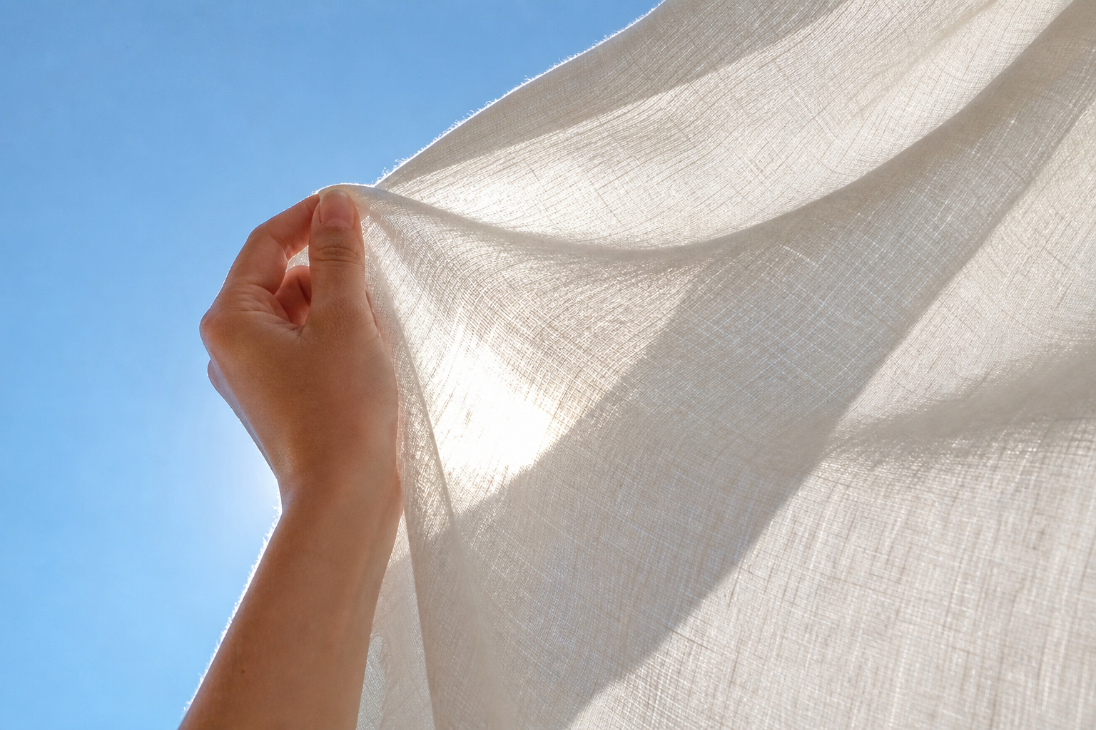 A hand holding a white linen curtain against sunlight and a clear blue sky.