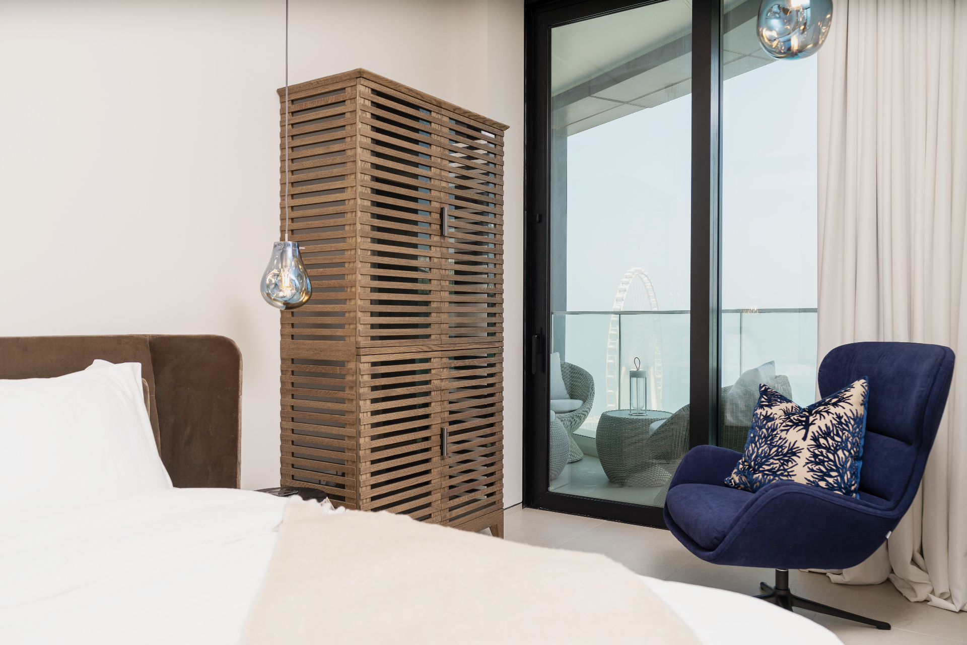 Primary bedroom detail — slatted oak wardrobe, navy velvet lounge chair and balcony access framing the sea and Ain Dubai
