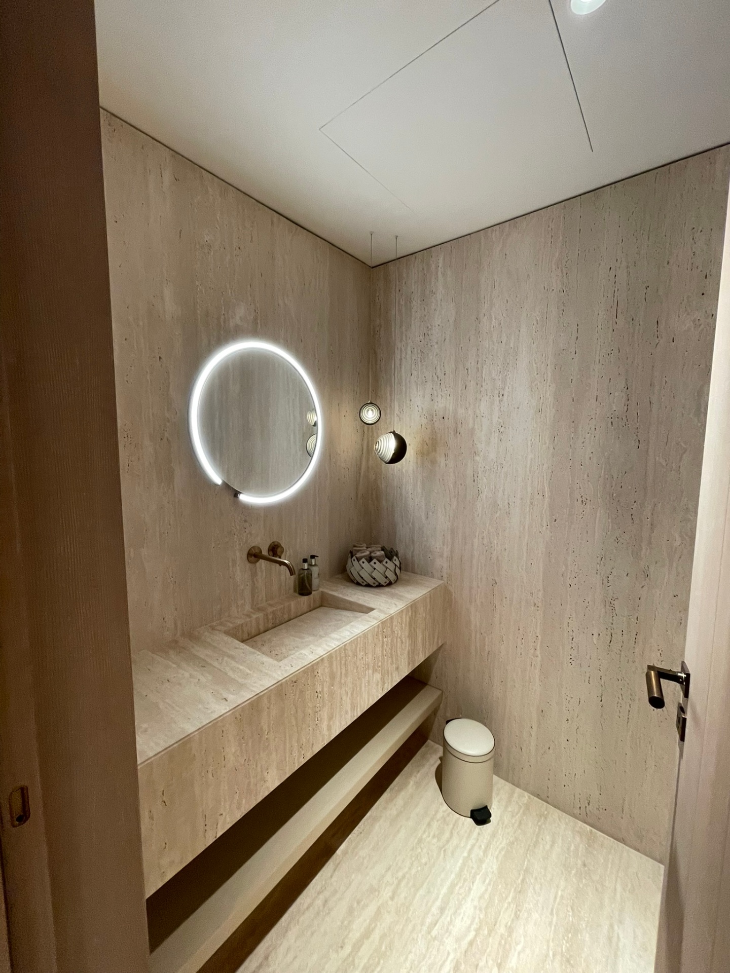 Powder room resolved in a single continuous plane of vein-cut travertine, with a backlit round mirror and suspended sculptural pendants