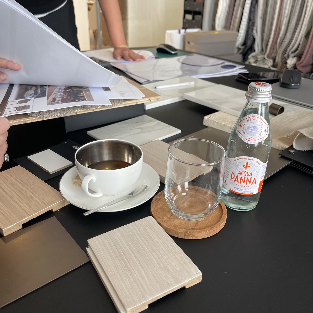 A designer turning drawings at a studio table set with timber, stone and metal samples, a coffee cup and a glass of water.